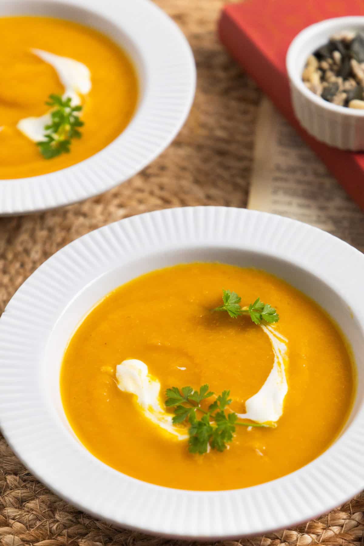 2 bowls of orange soup in a white bowl with a dollop of white cream and parsley as garnish and seeds in the background.