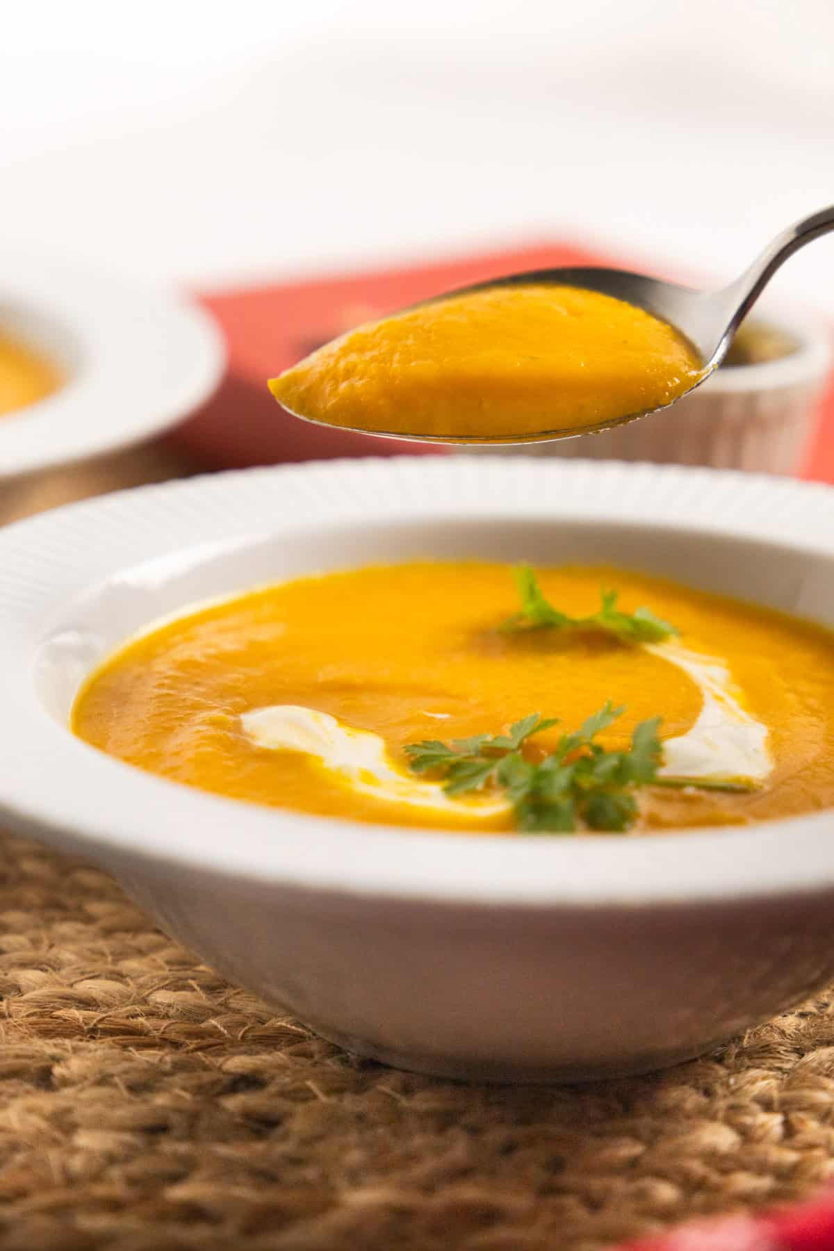 carrots soup in a white bowl with a dollop of white cream and parsley as garnish and a spoon showing the consistency. 