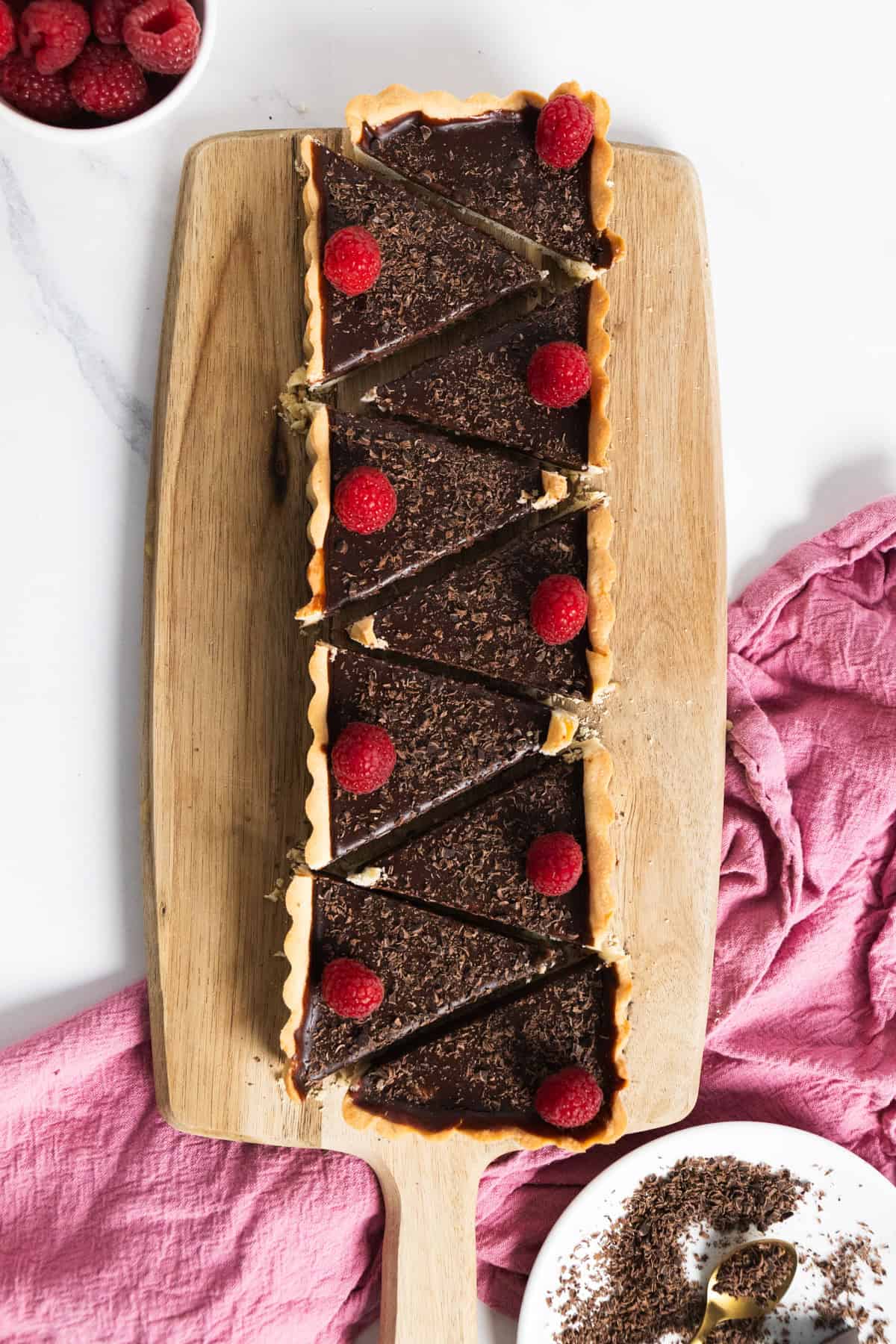 A rectangular chocolate tart on a wooden board cut in triangular slices with raspberries on each. 