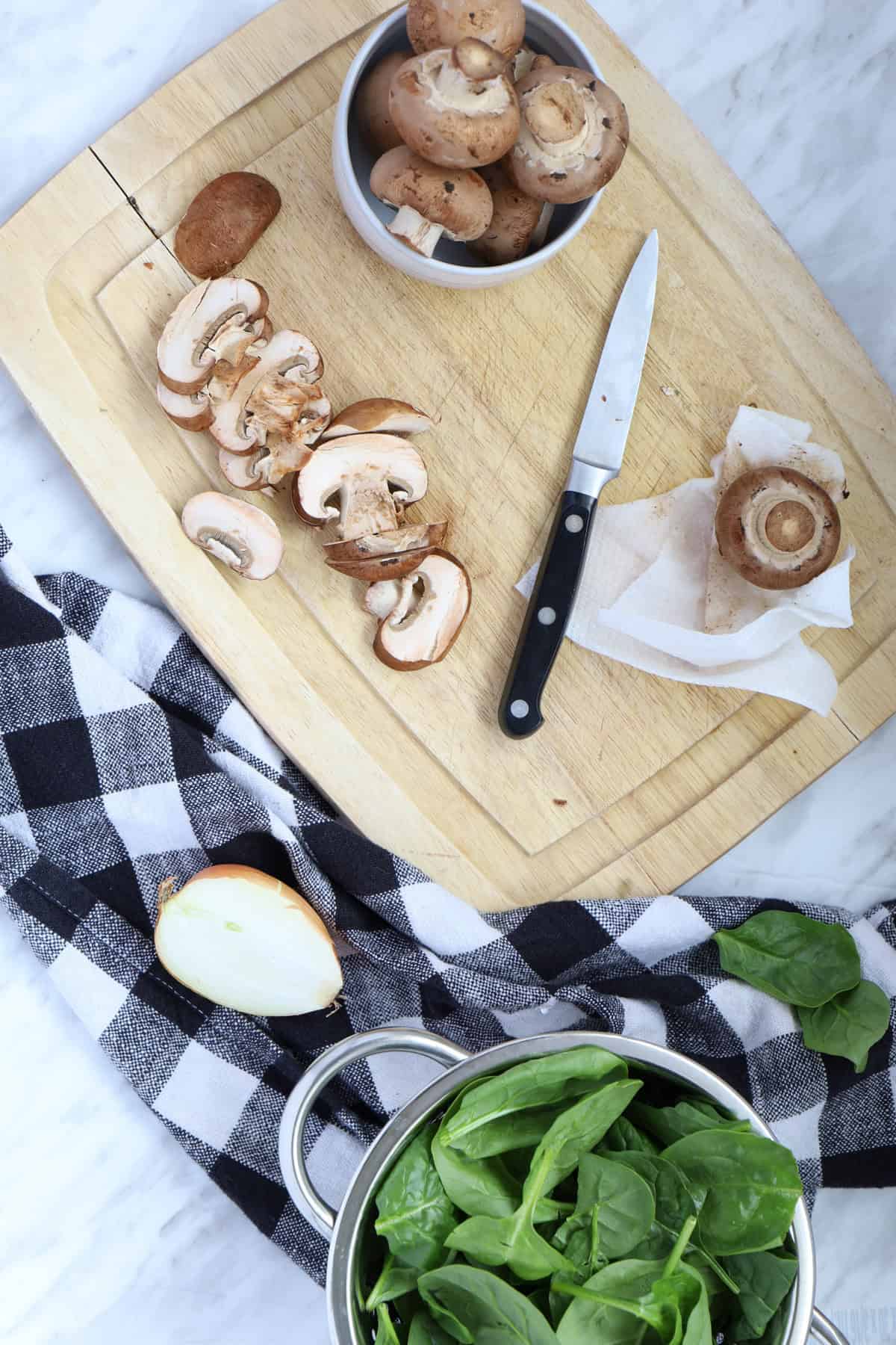 A wooden chopping board with sliced mushrooms and a knife. Spinach on the side.