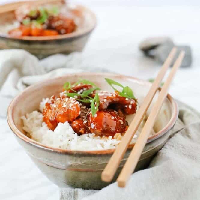 Side view of 2 brown bowls with sweet and sour chicken served over white rice, some chop sticks on top