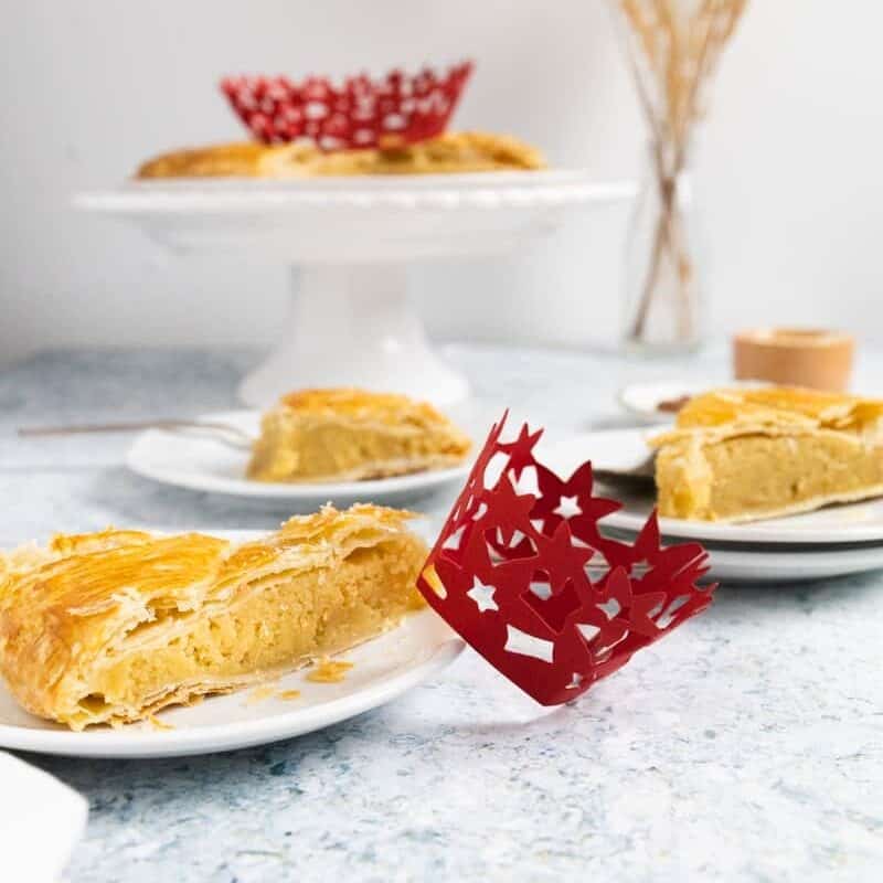 Side view of a plate with a slice of king cake with a mini red crown on the side and more slices in the bake and the rest of the cake on a cake stand.