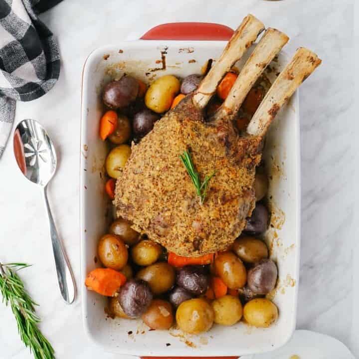 Top view of a rack of veal and potatoes in a rectangular dish, a spoon on the side, rosemary and honey